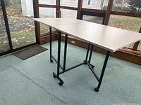 White folding table open with metal legs and caster wheels on carpeted floor with windows in background.