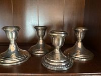 Four sterling silver weighted candlestick holders on wooden shelf, varying heights with one smaller Revere Silversmith piece and three Gorham pieces.