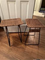 Photo of two Mid-Century Modern style stools with natural wicker seats and black metal legs, positioned on wood floor in front of white doors.