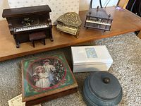 Three piano-shaped music boxes on a shelf with two decorative box-style and one metal cylinder style on floor