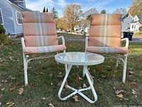 Front view of two white aluminum patio chairs with attached striped cushions and a small round glass table between them on grass.