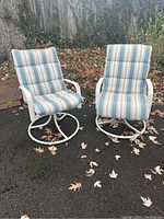 Front view of two patio swivel spring chairs with white metal frames and striped cushions on blacktop with leaf debris.