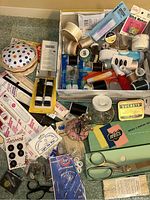 Lot photo of assorted sewing notions including pincushion, spools of thread, packaging for zippers, needle threaders, scissors, pins, button packages, and more.