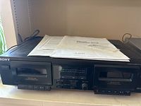 Front view of Sony stereo cassette deck with manual documents on top, showing dual cassette compartments and control panel.