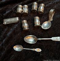 Photo showing six sterling silver salt and pepper shakers, three stainless spoons, and a napkin ring engraved Nancy, arranged on dark cloth