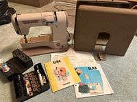 Full view of ELNA Supermatic sewing machine with storage case, accessory box, and manuals laid out showing included items.