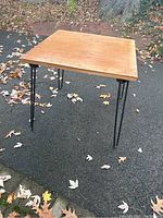 Front angled view of the vintage folding table showing the square solid wood top and black metal hairpin legs on asphalt with dry leaves around.