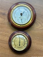Top image shows two vintage wall weather gauges mounted on a wood surface, barometer on top and hygrometer below, both round with wood frames and brass edges.