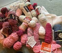 Overview photo showing multiple skeins and balls of yarn in various colors such as pink, red, white, and blue, placed on a carpeted floor.