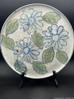 Front view of the round ceramic plate with blue and green floral decorations on off-white speckled base.
