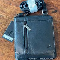 Front view of black leather crossbody purse with attached tags and adjustable strap coiled at top.