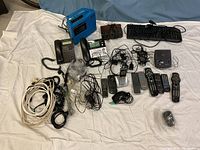 Wide view showing all electronics including vintage Panasonic tape deck, phones, remotes, keyboard, and cords on a white sheet.