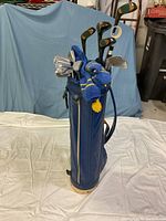 Full view of the blue vintage golf bag with golf clubs inside, showing the club heads and partial bag structure, background blue drape.