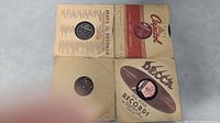 Four vintage 78 RPM records in their original paper sleeves. One sleeve shows Decca Records, one Columbia, one Bluebird, and one Capitol. All have visible labels on the records.