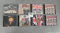 Photo showing all 8 CD jewel cases laid flat on gray surface, includes albums by The Hollies, Herman's Hermits, Robert Plant, and British rock compilations.