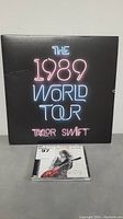 Black album cover with neon style text '1989 World Tour Taylor Swift' behind a CD case.