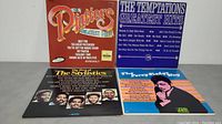 Four vinyl record albums standing upright on a gray surface: The Platters Greatest Hits (red cover), The Temptations Greatest Hits (blue cover), The Stylistics album (black cover with members' photos), Percy Sledge album (striped orange and pink cover).