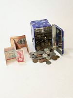 Coins spilling out of a small decorative blue box with white flowers, next to folded paper currency including a bill with Queen Elizabeth II portrait.