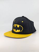 Front view of the snapback hat showing embroidered Batman logo in yellow on black crown and yellow brim.