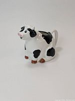 Front-side view of the white ceramic cow pitcher showing black spots, pink snout, brown hooves, and white handle.