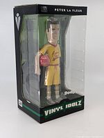 Photo of vinyl figure in original black and green Vinyl Idolz box with clear plastic front window, showing figure holding dodgeball with 'Average Joes' uniform.