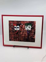 Front view of the framed print featuring cartoon Joker and Harley Quinn characters against a brick wall background. The print is double matted in white and enclosed in a red frame, displayed on a stand.