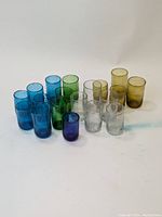 Full lot of 19 handblown glass cups arranged by color: light blue, dark blue, green, clear, yellow.