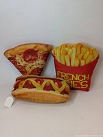 Full view of the three throw pillows styled as food items: pepperoni pizza slice, hot dog with mustard, and red container of French fries with yellow text.