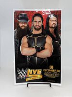 Front view of the laminated WWE tour poster showing Roman Reigns prominently in center with two other wrestlers. The WWE Live Holiday Tour logo and event date appear at bottom.