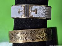 Photo showing front view of two bracelets: white leather with gold-tone rhinestone decorated clasp and brass engraved floral bracelet.