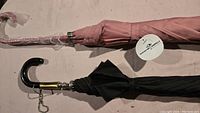Two umbrellas laid out flat, one pink wrapped in plastic, the other black with curved handle