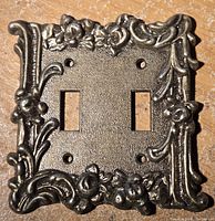 Front view of the brass double light switch cover showing ornate floral and scroll detailing around edges.