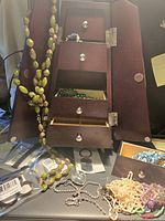 Brown wooden jewelry box shown open with four drawers and two side hinged doors. Contains assorted costume jewelry such as green beaded necklace, silver rhinestone chain, white beaded necklace, and other pieces.