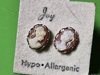 Close-up photo of pair of cameo post earrings showing white cameo female profiles on burgundy background set in silver-tone frames on a card marked 'Joy' and Hypo-Allergenic.