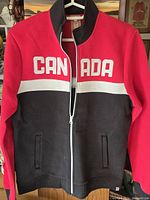Front view of women's jacket sweater showing 'CANADA' spelled out across chest in white letters, color blocked in red (upper), white (stripe), and black (lower).