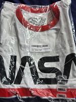 Front view of folded white Tee Luv NASA Classic T-Shirt with red collar inside clear plastic packaging showing partial NASA logo and size small sticker