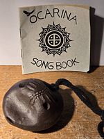 Photo of the handmade ocarina placed in front of the ocarina songbook showing the full booklet cover and the raised feather design on the instrument.