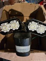 Two photos showing the outdoor security light unit with dual LED floodlight heads and central motion sensor from top down and angled perspective in cardboard box.