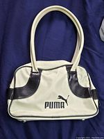 Front view of a cream-colored Puma purse with black leather accents and Puma logo on front, placed on a dark blue background.