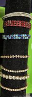 Seven rhinestone bracelets on a black display stand showing variety in colors and design including clear, red, and blue rhinestones