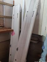 Two white metal security scissor gate panels leaning against a wall, showing vertical bar design, lock housings, and mounting points.