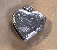 Close-up front view of a polished sterling silver heart-shaped locket with floral engraving and a small loop for attaching to a chain.