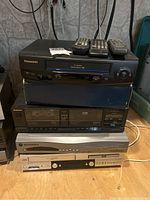 Stacked electronic devices including a Panasonic VHS player, Technics stereo cassette deck, Motorola StarChoice receiver, and JVC VHS/DVD player.