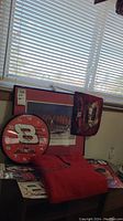 Dale Earnhardt Jr. themed clock, framed print, red flag, and folded red hoodie displayed on floor near window.