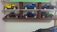 Six diecast model cars placed on two wooden shelves. Top shelf has three cars: yellow 1932 V8 Convertible Sedan, blue 1934 V8 Deluxe Roadster, dark 1939 Ford Deluxe Convertible Coupe. Bottom shelf has three cars: black 1940 Ford Deluxe Business Coupe, dark 1933 Chevrolet Two Passenger Five Window Coupe, green 1930 Chevrolet Coach.