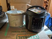 Granitestone multi-cooker with manual booklet on top and Proctor-Silex Crock Watcher slow cooker placed on a box.