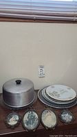 Photo showing metal cake plate with aluminum dome, stacked wedding anniversary plates, and four small framed prints lined in front.
