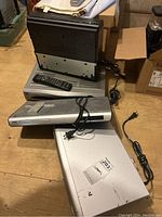 Photo showing three devices stacked on a wooden table: an RCA DRC279 DVD player with remote on top, a silver Panasonic VHS player, and a Motorola device below. Power cords are visible.