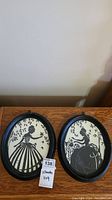Pair of two black oval silhouette framed artworks placed on wooden table, showing full frames and art inside