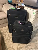 Photo showing two black rolling suitcases on carpet, one larger behind smaller with emblems visible